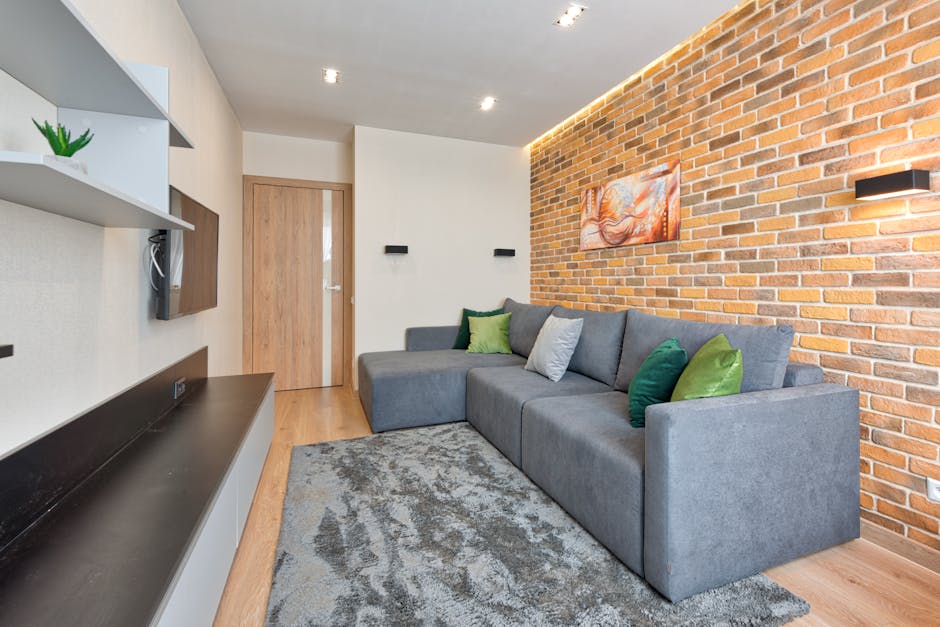 A modern living room featuring a large L-shaped grey fabric sofa with six decorative cushions in shades of green, grey, and white, positioned against a brick accent wall with warm orange tones. The room has wooden flooring with a textured grey area rug in front of the sofa. On the left, a wall-mounted white shelving unit displays a small green potted plant, while a black shelf below holds a TV. The space is illuminated by recessed ceiling lights and a sleek black wall sconce on the brick wall, contributing to a clean and tidy appearance suitable for deep surface cleaning and sanitisation by Lambeth Carpet Cleaners.