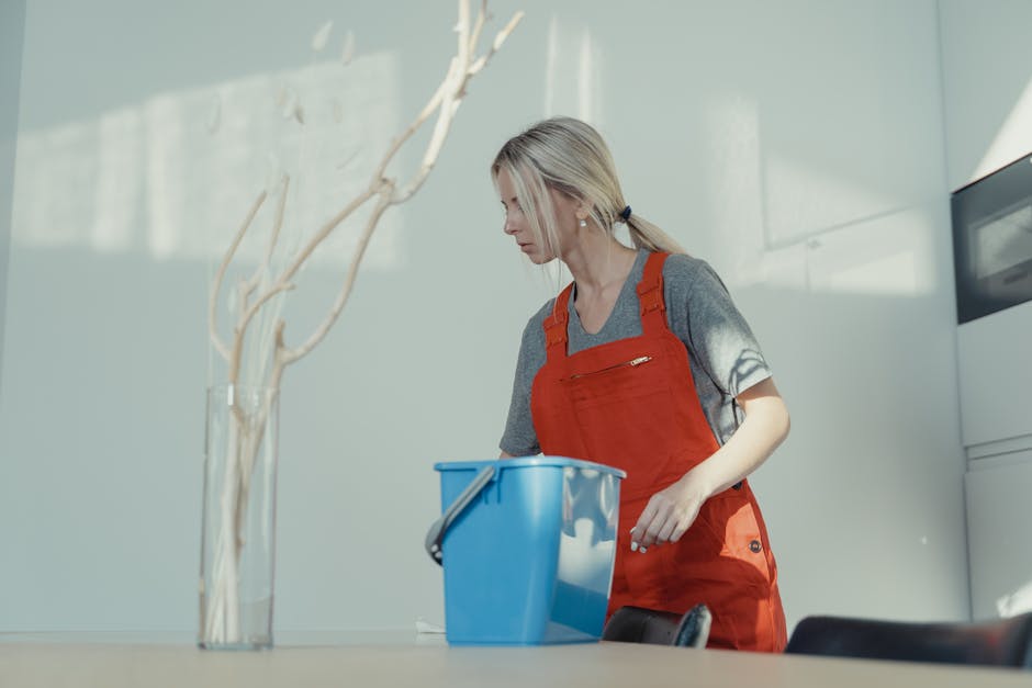 Two professional cleaning staff members from Lambeth Carpet Cleaners standing back-to-back against a plain white wall in a well-lit room. The woman on the left, of Asian descent, is holding a vacuum c