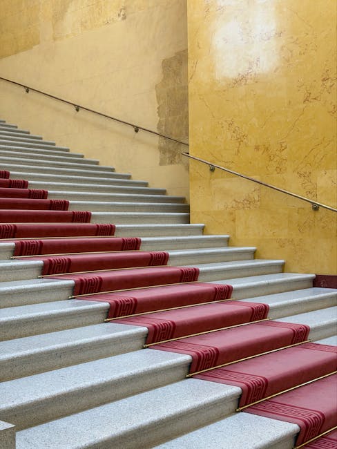 A set of indoor marble stairs with a polished, smooth surface, featuring a red carpet runner with a geometric pattern along the center. The stairs are bordered by a brass trim and lead up to a landing