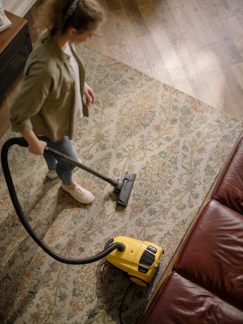 A woman is using a yellow vacuum cleaner with a black hose on a large, patterned area rug that features floral designs in soft pastel colors. She is standing on the rug in a living room, with wooden flooring visible at the edges and a red leather sofa nearby. The room is well-lit, with natural light illuminating the space, and the surface appears clean and dry, suggesting a deep cleaning or surface sanitisation process. Lambeth Carpet Cleaners specializes in domestic cleaning services, including surface cleaning and hygiene maintenance, as seen in this detailed carpet cleaning scene.